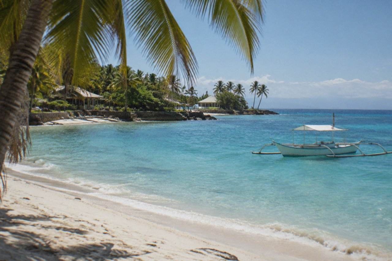 A palm-fringed white sand beach on Mactan Island with calm turquoise water and a small outrigger boat anchored offshore