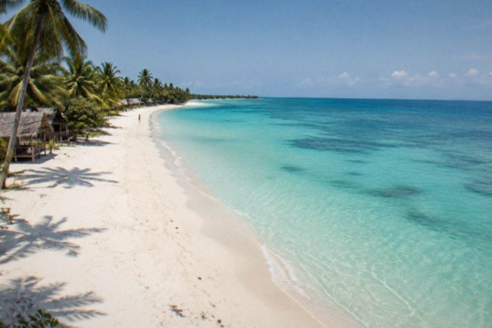 A wide empty white sand beach on Bantayan Island at midday with a few native-style cottages under coconut palms along the shoreline