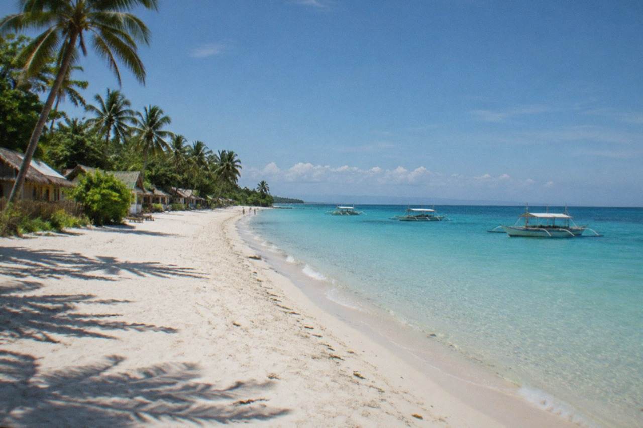 A wide white sand beach on Bantayan Island in Cebu, Philippines, with calm turquoise shallows and a few bancas moored offshore