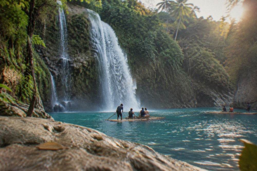 Swimmers on bamboo rafts in the blue pool below the main cascade at Kawasan Falls, with jungle cliffs rising on both sides