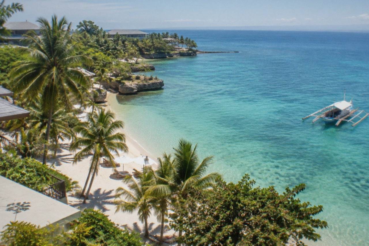 A resort beach on Mactan Island with calm turquoise water and a wooden outrigger boat moored offshore