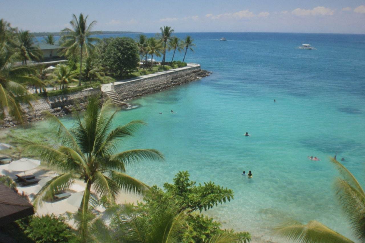 A calm turquoise swimming area along a Mactan beach at midday with a low stone seawall and palm trees