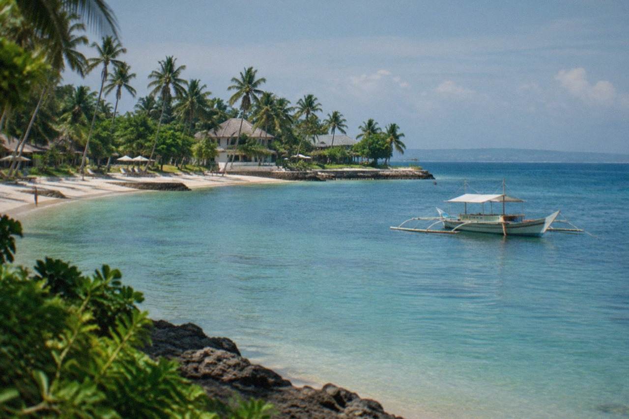 A resort beach on Mactan Island with calm turquoise water and a wooden outrigger boat anchored offshore