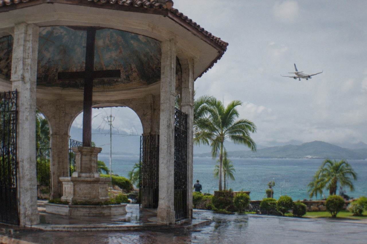 A passenger plane descending over coastal waters toward an island airport with green hills in the background