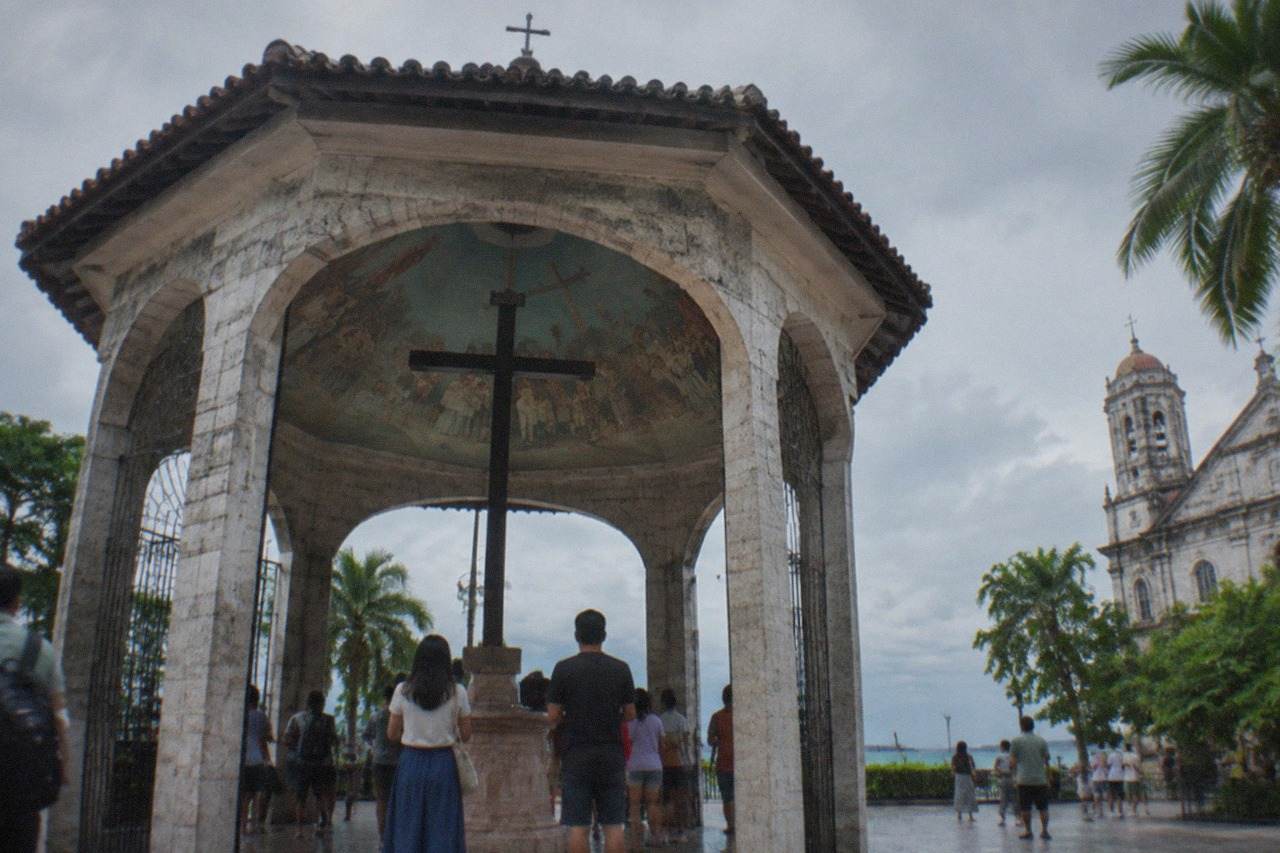 A painted wooden cross housed in an open stone pavilion, with visitors gathered around it and an old church street beyond