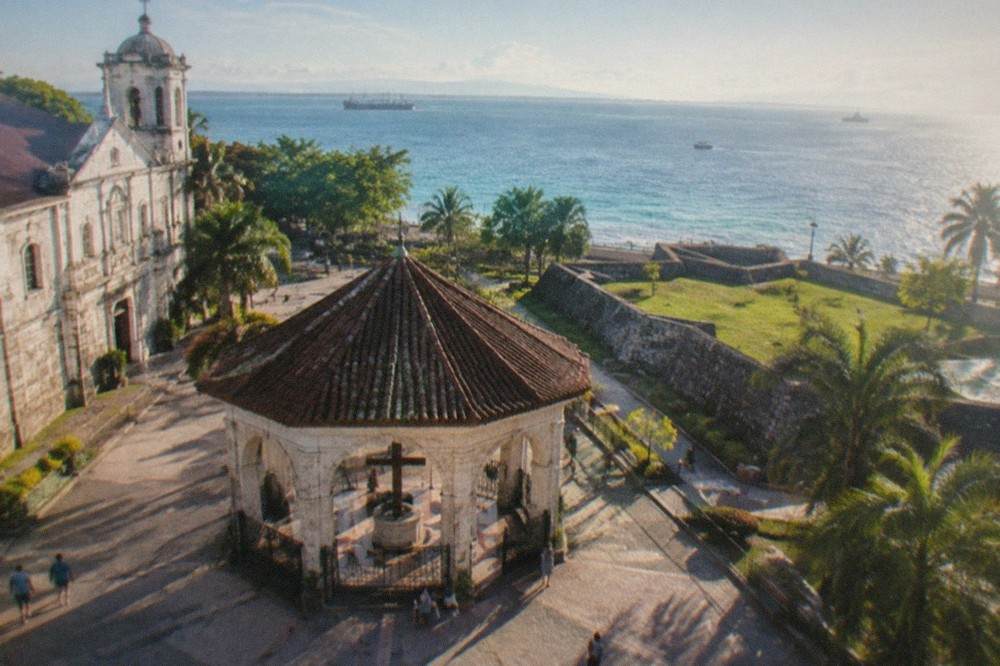 A low stone fort with triangular walls and a grassy courtyard, facing a plaza near the sea