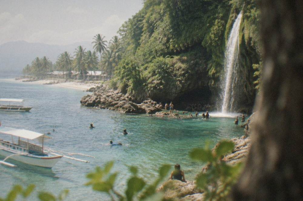 Tall thin waterfall pouring over a green mossy limestone cliff into a shallow natural pool surrounded by visitors