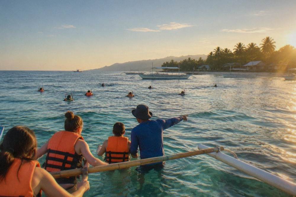 Visitors in life jackets holding on to a bamboo outrigger boat while a guide points toward the water