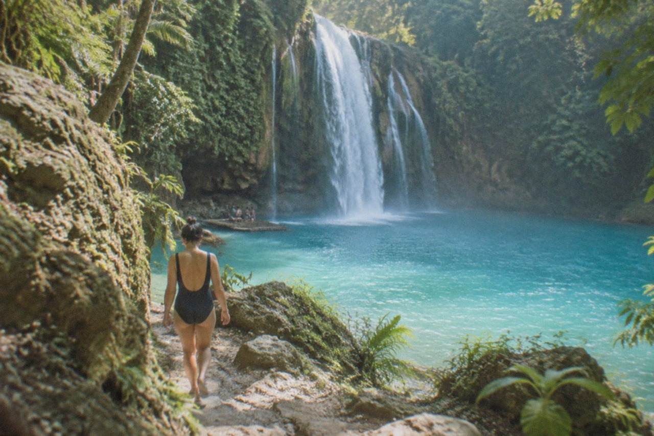 Kawasan Falls in Badian, Cebu, with turquoise water pooling under a jungle cliff and a small wooden raft at the base of the falls
