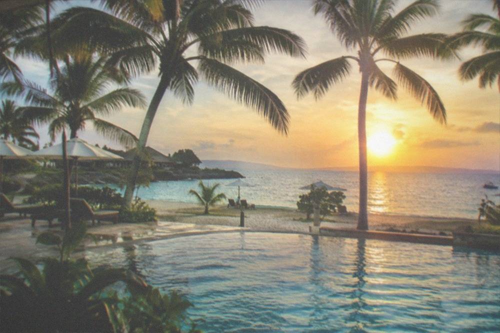 A palm-fringed resort swimming pool beside a beach at sunset