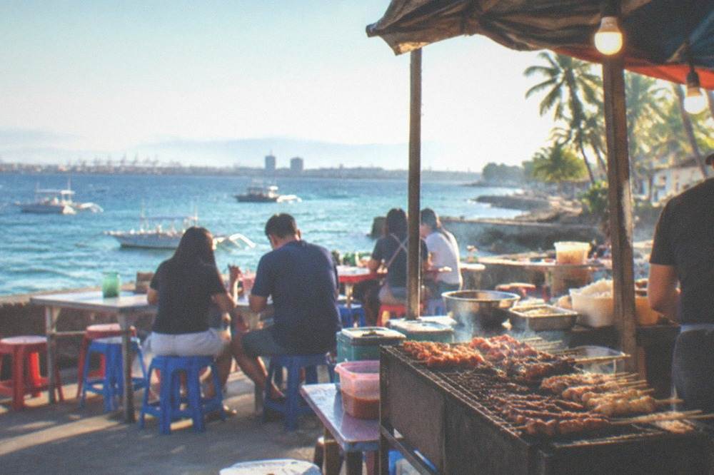 Small food stall serving grilled skewers and rice dishes at night with plastic stools and warm lighting