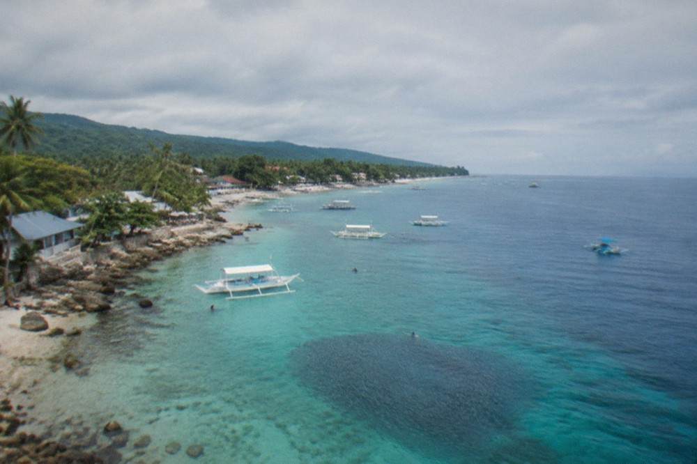 Aerial view of a rocky beach strip with boats near turquoise water and green hills behind