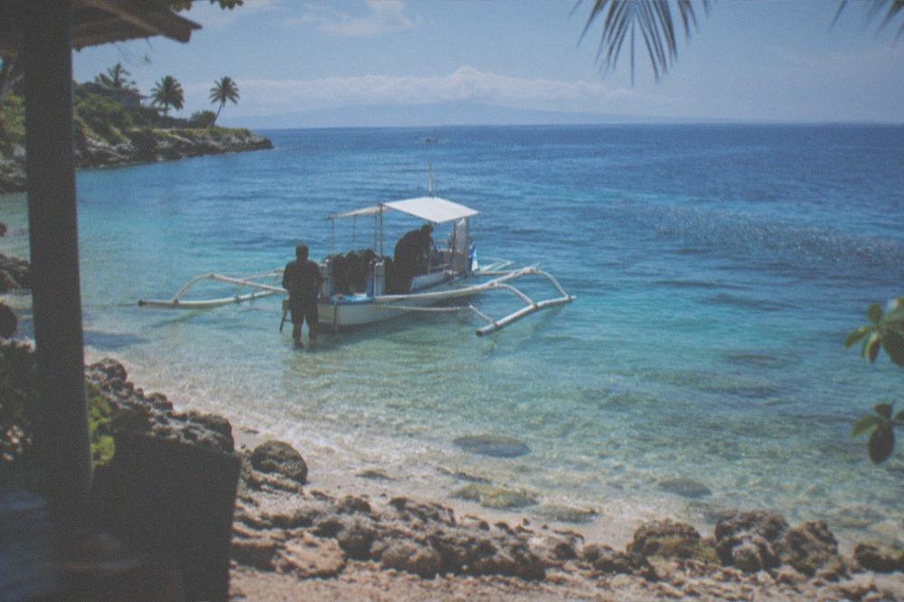 A small outrigger boat moored in shallow clear water with divers loading gear