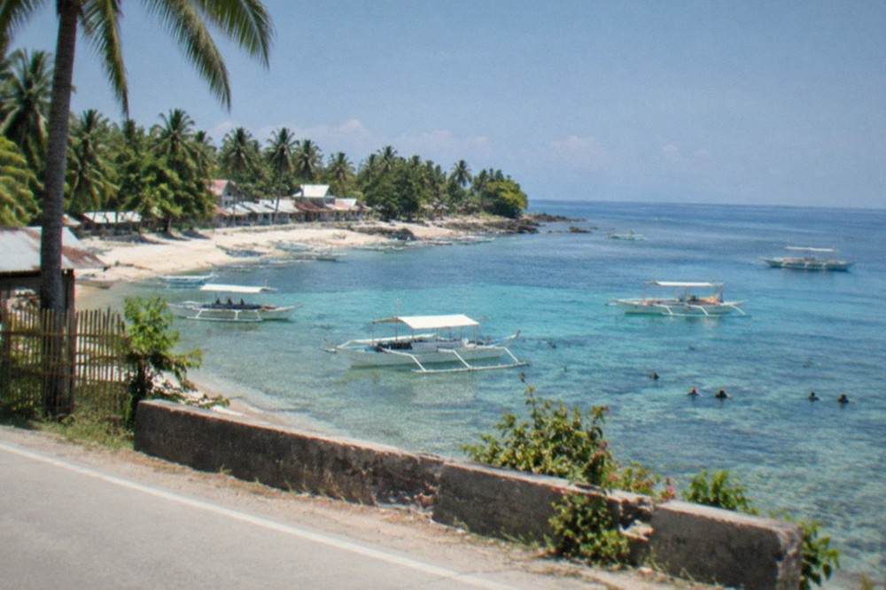 A small coastal Philippine town seen from a road, with low buildings, palm trees and a calm blue sea beyond