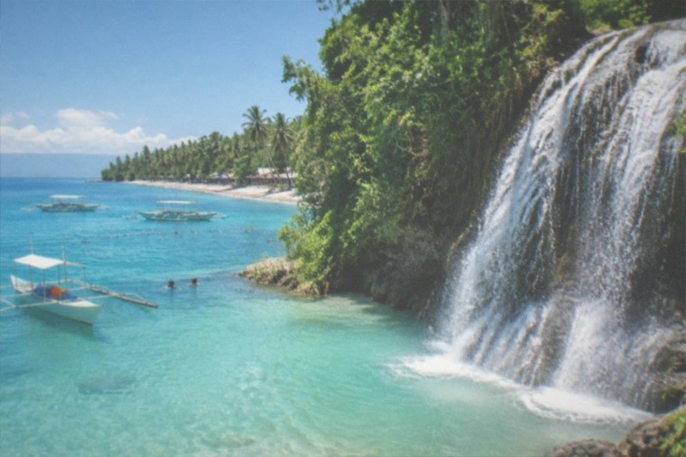 A wide waterfall fanning over dark rock into a shallow turquoise pool ringed by tropical plants