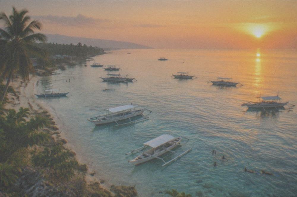 A calm tropical beach at sunrise with soft orange light over the water and silhouetted boats