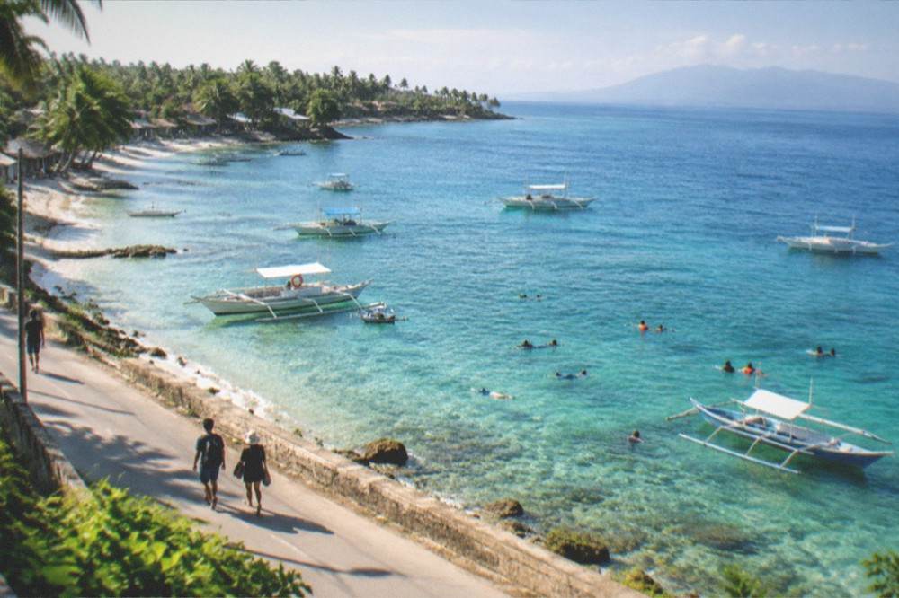 Travellers walking on a coastal road at dawn with fishing boats moored in shallow water nearby
