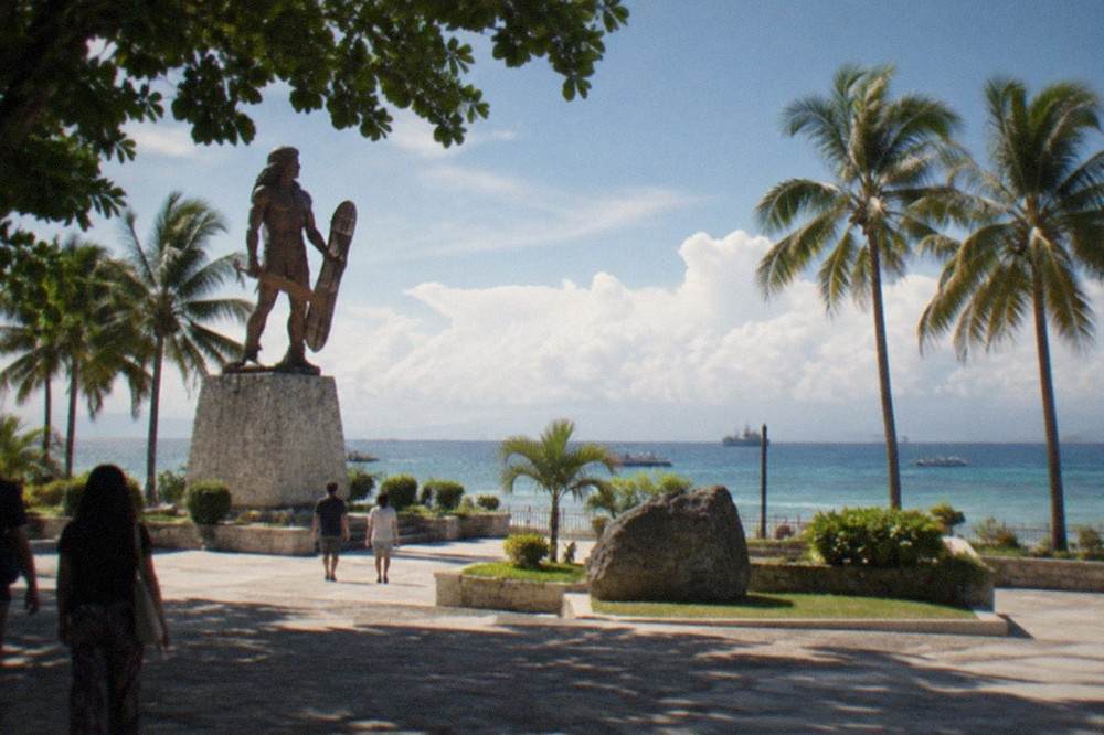 A wide plaza with palm trees and a stone mural wall beside the sea, a few visitors walking in the shade