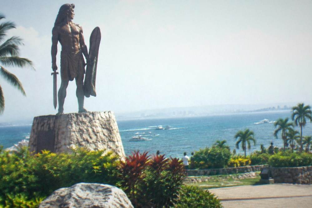 A stone mural wall showing carved scenes of warriors fighting on a beach, with tropical planting in front