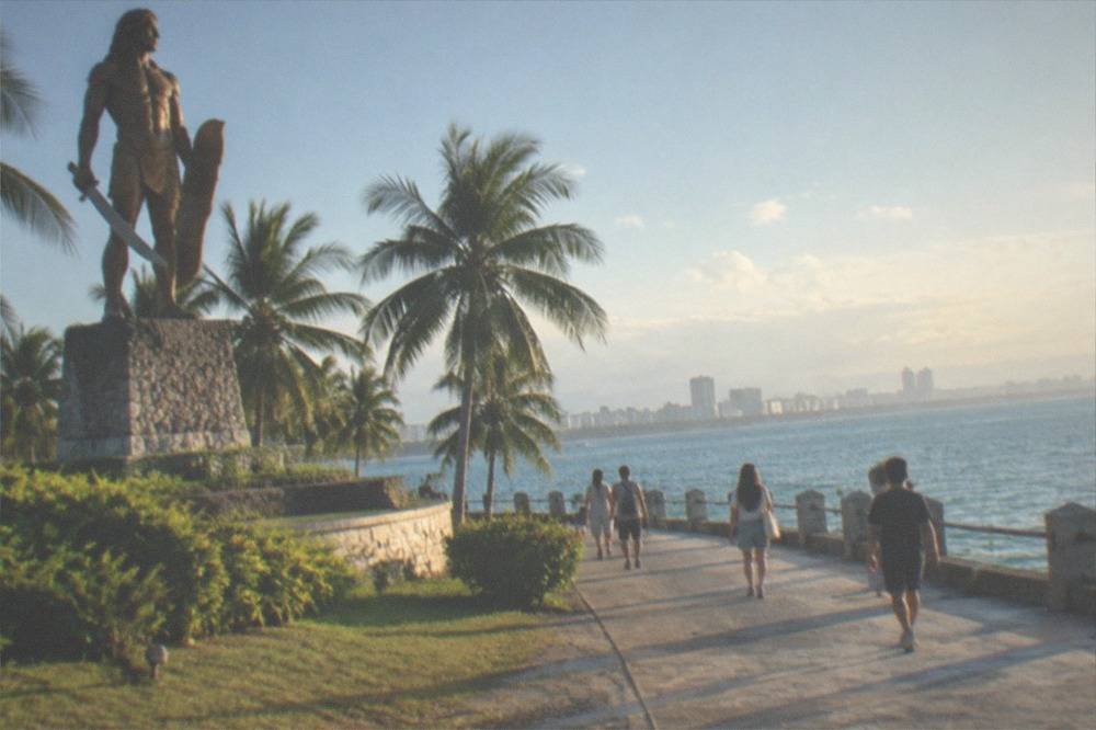Visitors walking along a paved seafront promenade with palm trees and a view across a channel to a city skyline