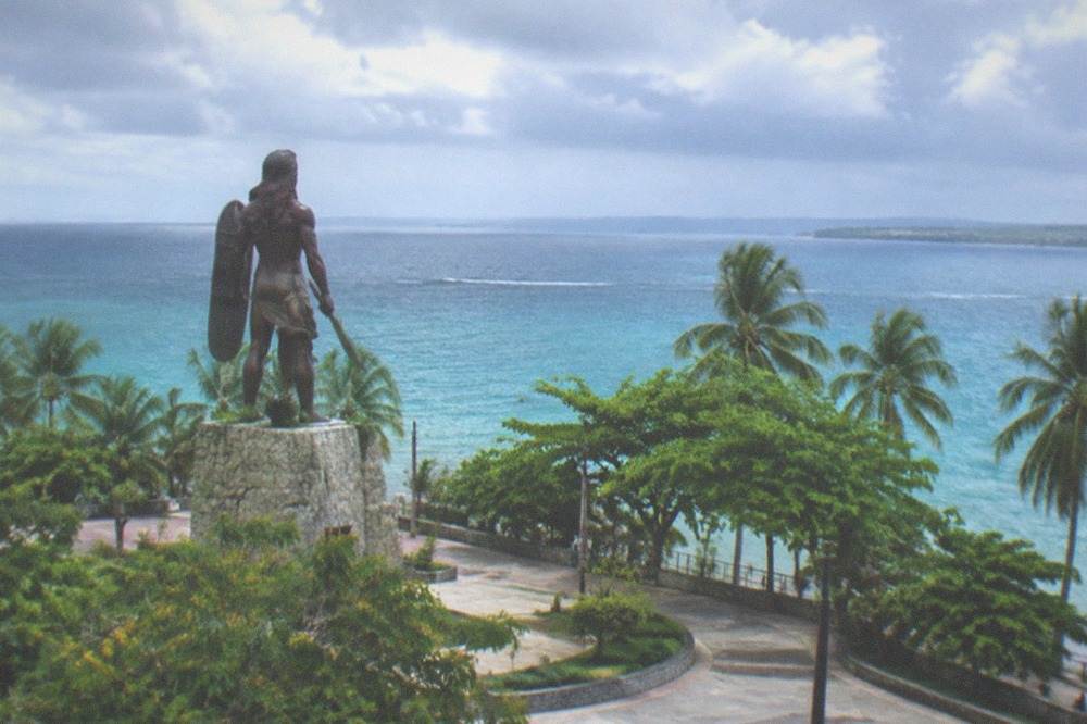 A shaded plaza with mature trees, commemorative plaques on a low wall and a glimpse of the sea beyond