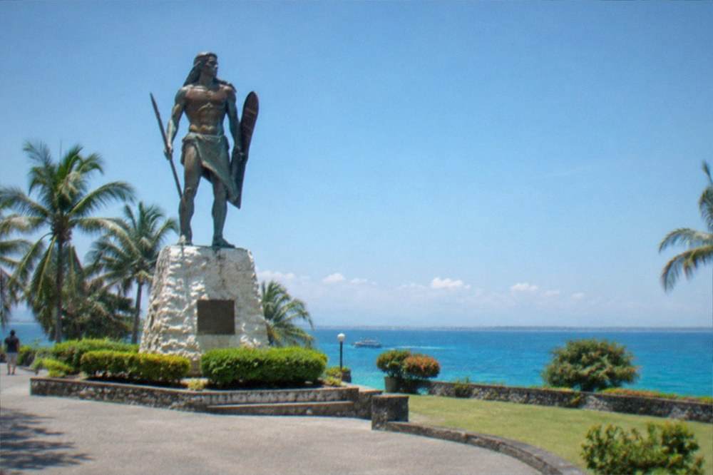 A bronze statue of a chieftain with a raised bolo silhouetted against a bright morning sky over the sea