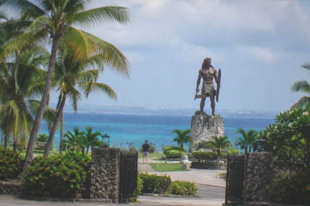 A park gate with tropical planting and a view of a tall statue in the distance under a partly cloudy sky