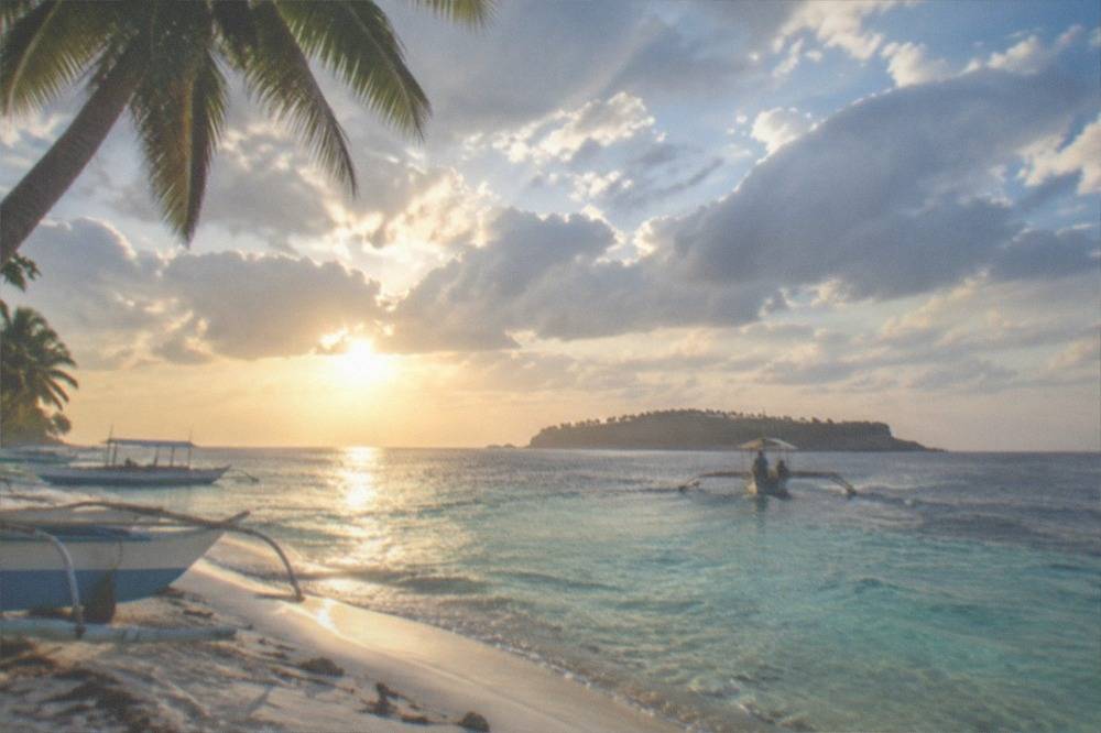 A wooden outrigger boat crossing open water toward an island at sunrise