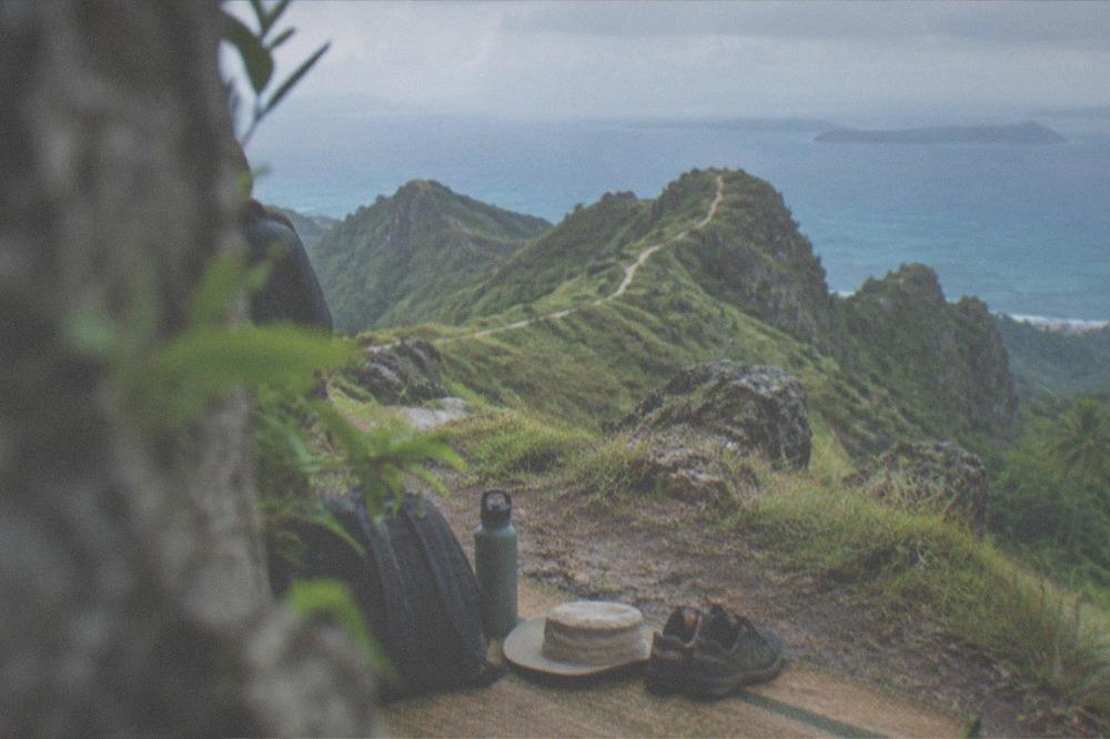 A small backpack, water bottle, hat and walking shoes laid out on a woven mat before a hike