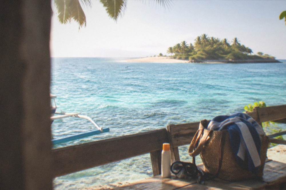 A beach bag with a towel, snorkel mask and sunscreen on a wooden bench by the sea