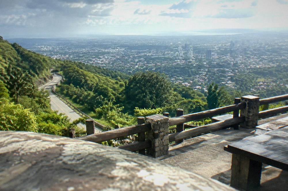 Winding mountain road climbing through green tropical hills with a city visible far below