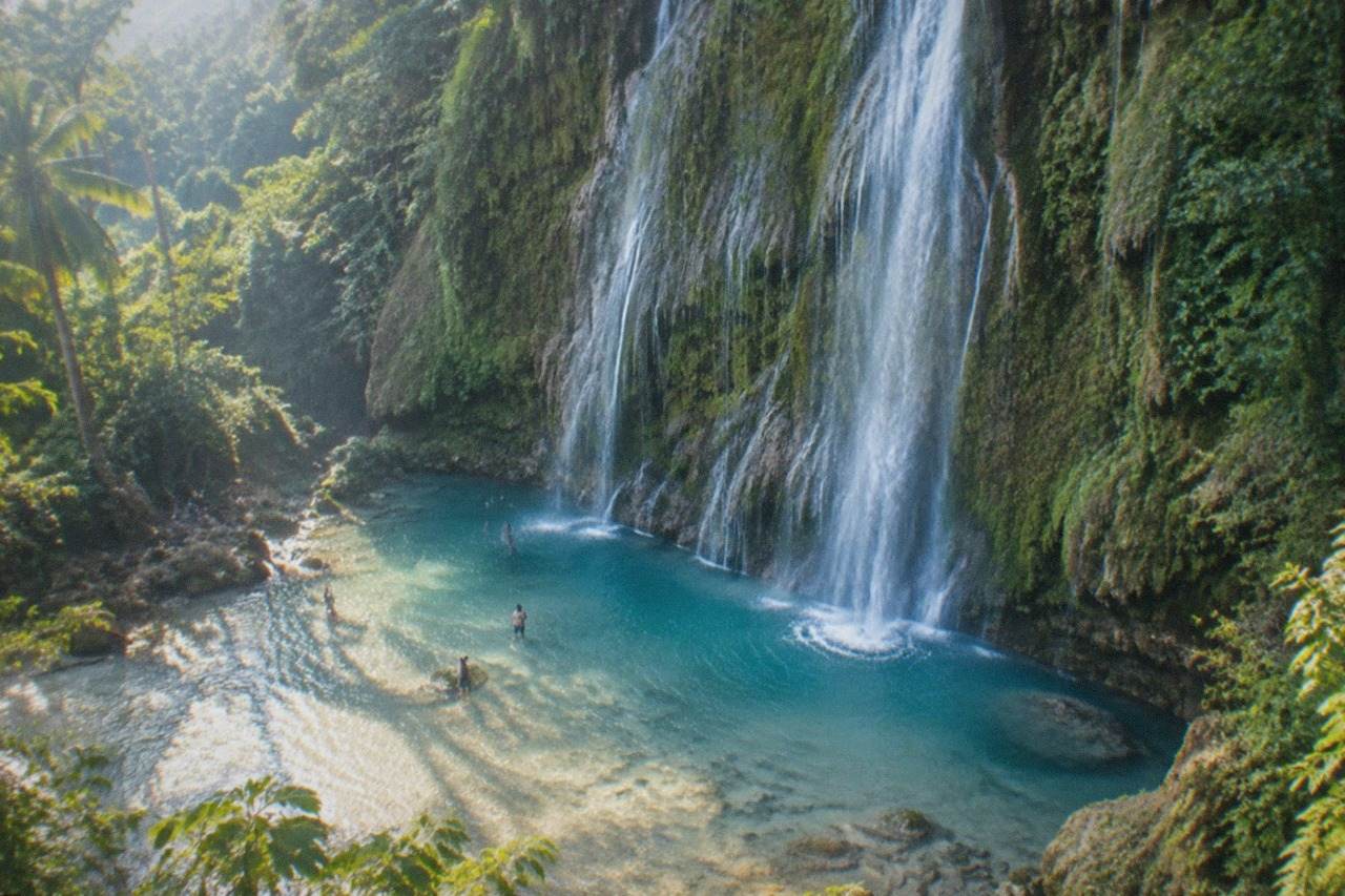 A tall thin waterfall pouring over a mossy limestone cliff into a shallow turquoise pool