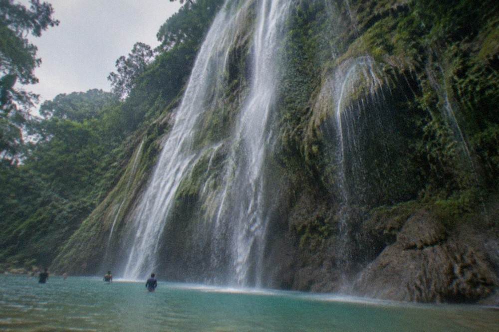 Visitors wading in the shallow pool beneath a wide dripping rock face at a tropical waterfall