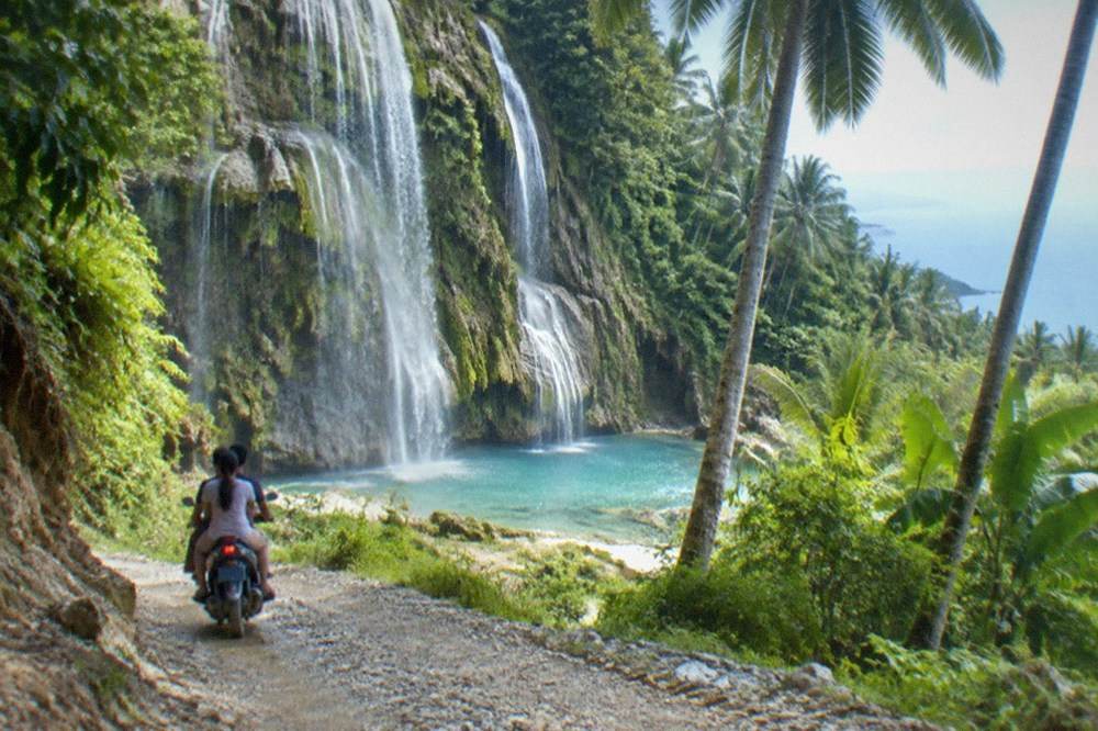 A motorbike with two passengers descending a steep gravel track through coconut palms