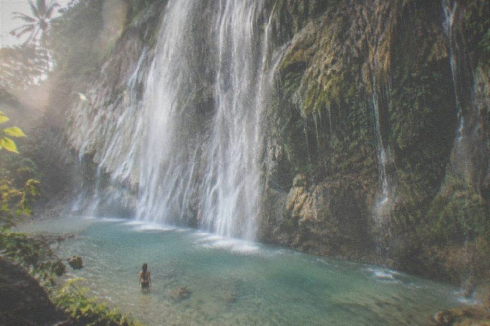 A person standing waist deep in a shallow pool looking up at a wide waterfall