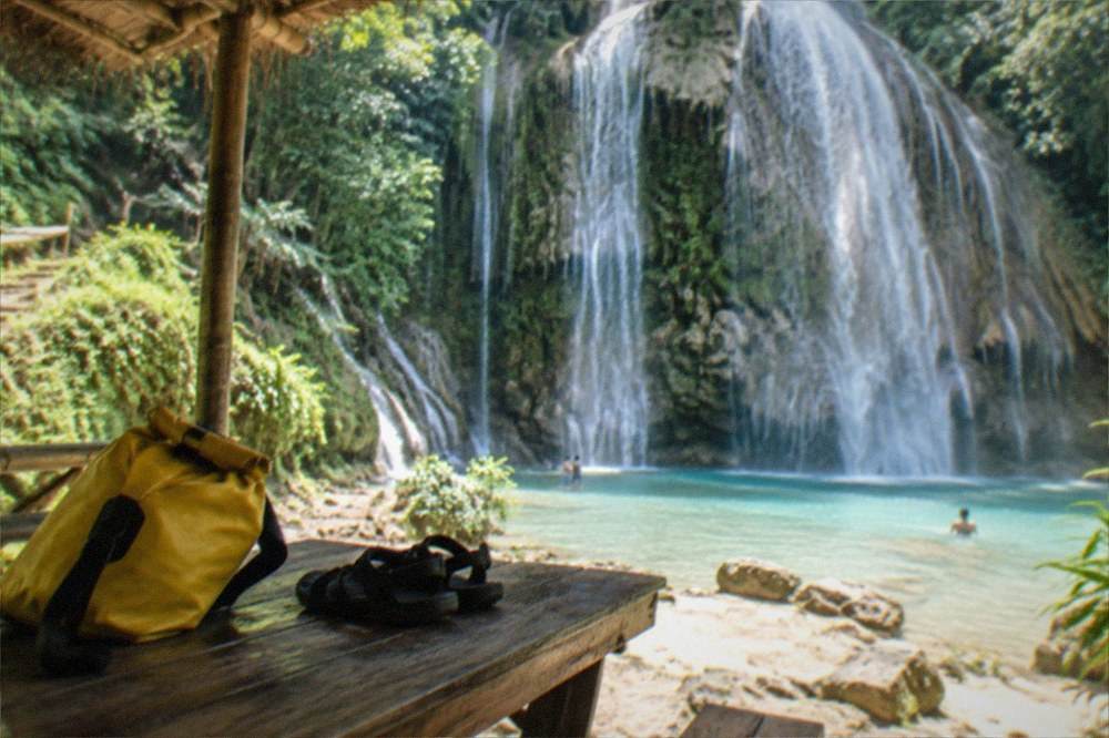 A dry bag and sandals resting on a picnic table under a simple bamboo shelter near a waterfall