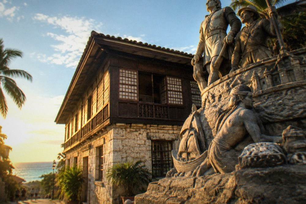 A sculpted monument depicting scenes from Cebu's history, viewed from a narrow street in the Parian district