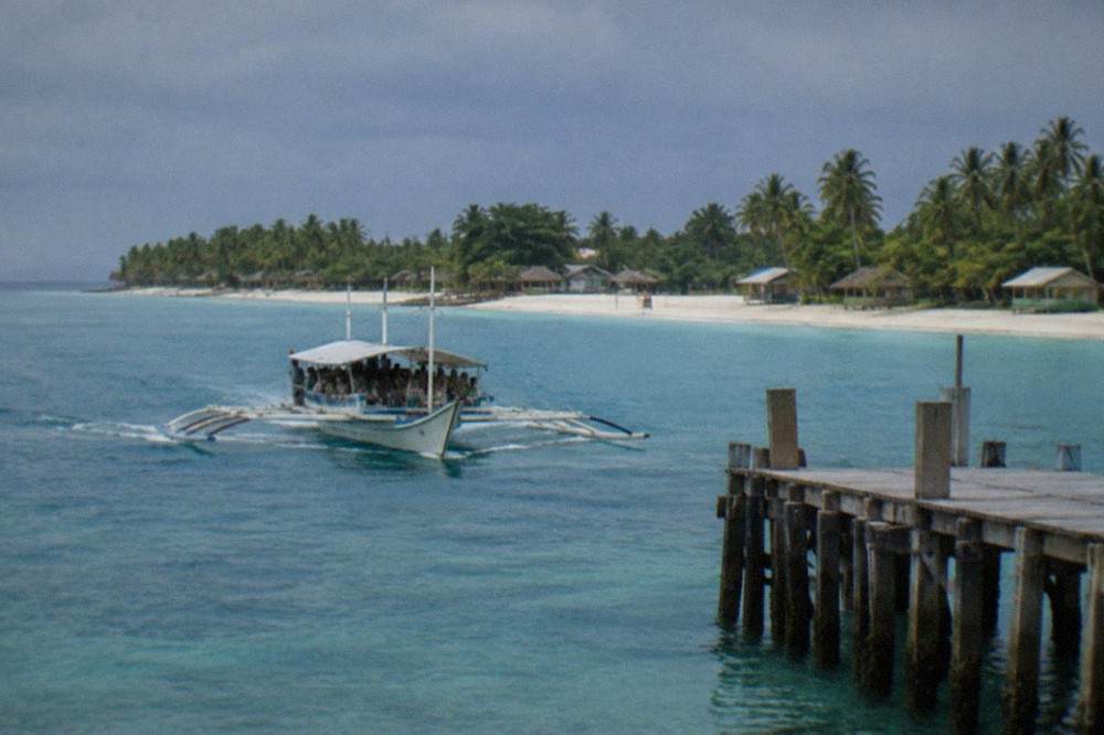 A small outrigger ferry loaded with passengers approaching a wooden pier on a tropical island