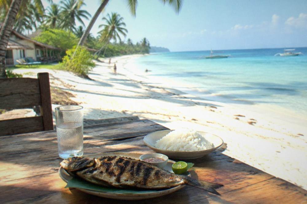A grilled fish and rice lunch served on a wooden table overlooking a white sand beach