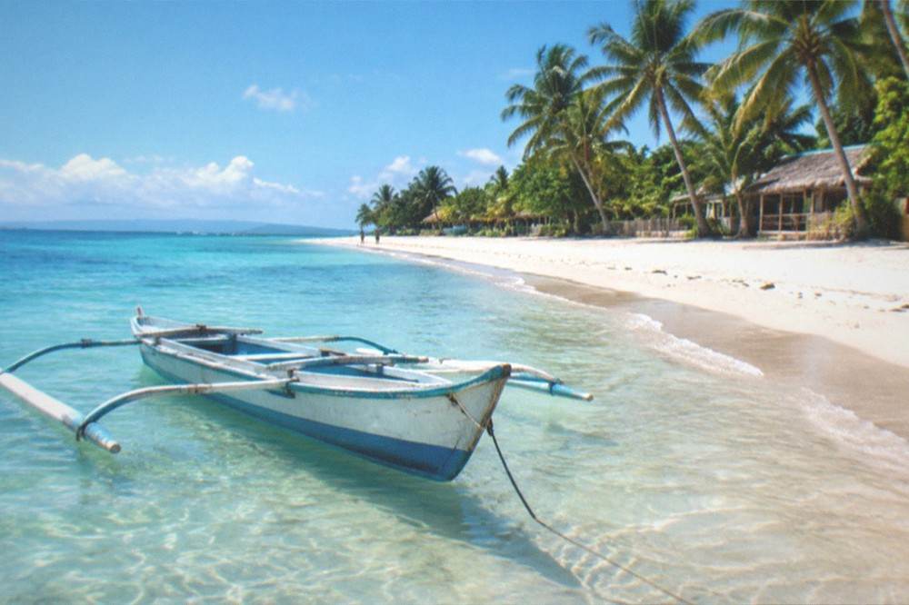 A quiet stretch of white sand beach with a small outrigger boat resting in shallow clear water