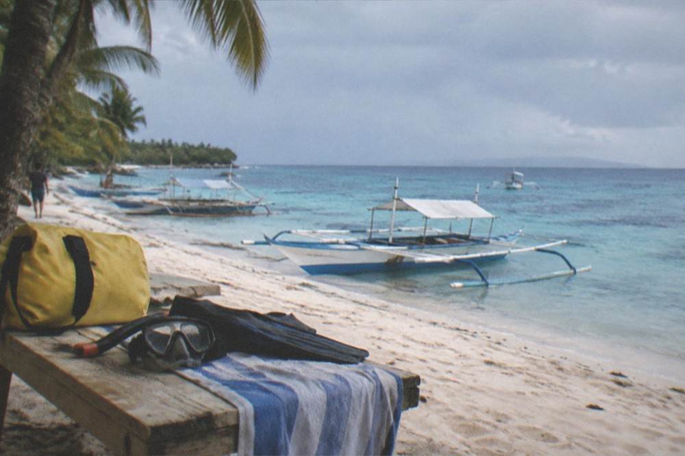 A canvas dry bag and snorkel gear laid out on a wooden bench beside a beach towel