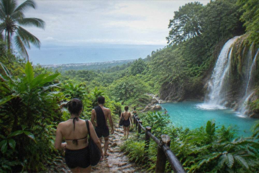 Travellers in swim gear walking down a jungle trail toward a turquoise waterfall pool in south Cebu