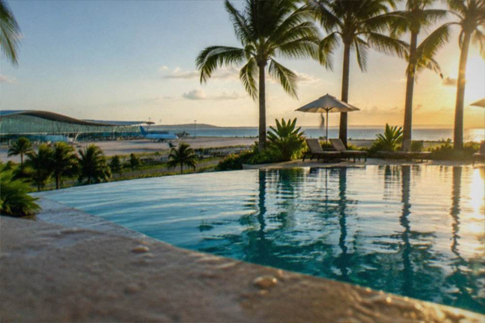 A resort swimming pool with palm trees and lounge chairs facing the sea at sunset
