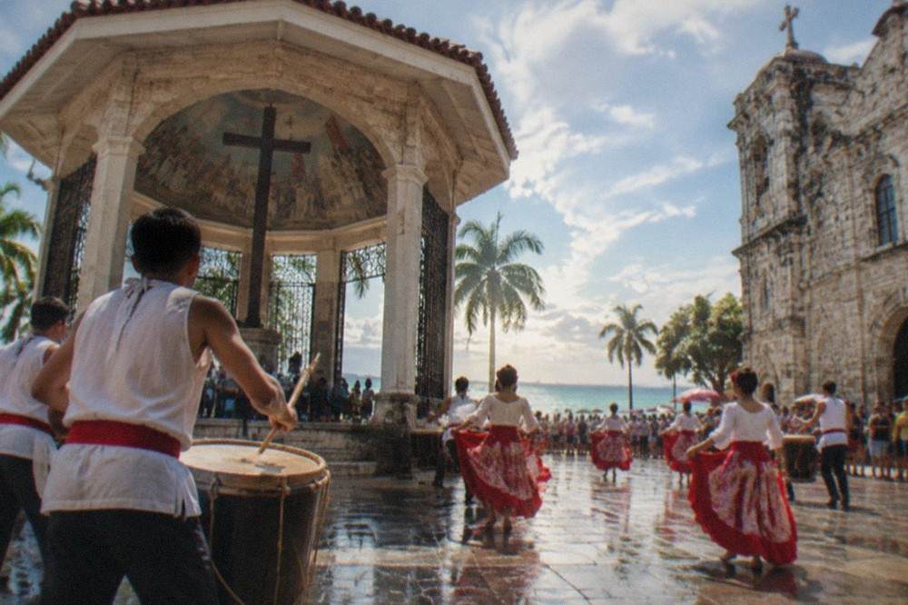Drummers and dancers moving through a crowded plaza beside a historic stone church facade