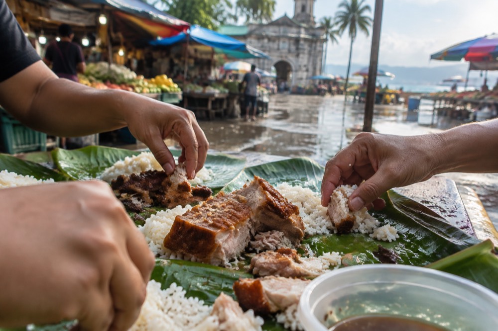 Hands sharing roast pork and rice from banana leaves spread across a table