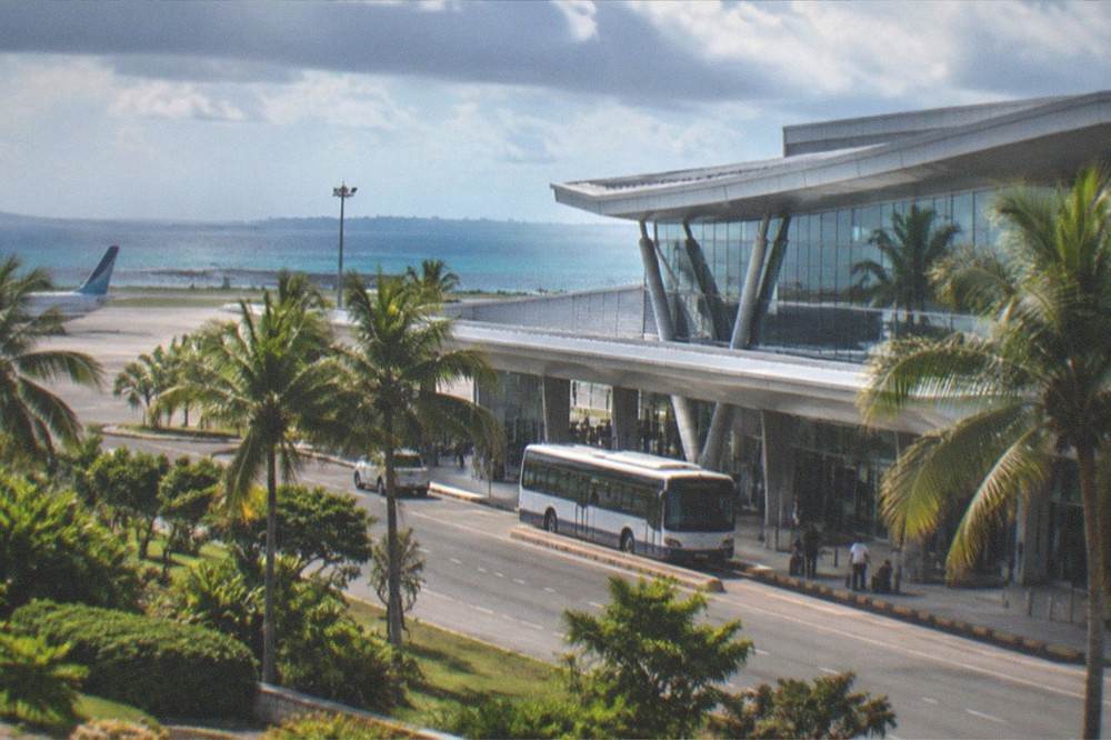 A modern airport terminal exterior with palm trees and a shuttle bus at the kerb