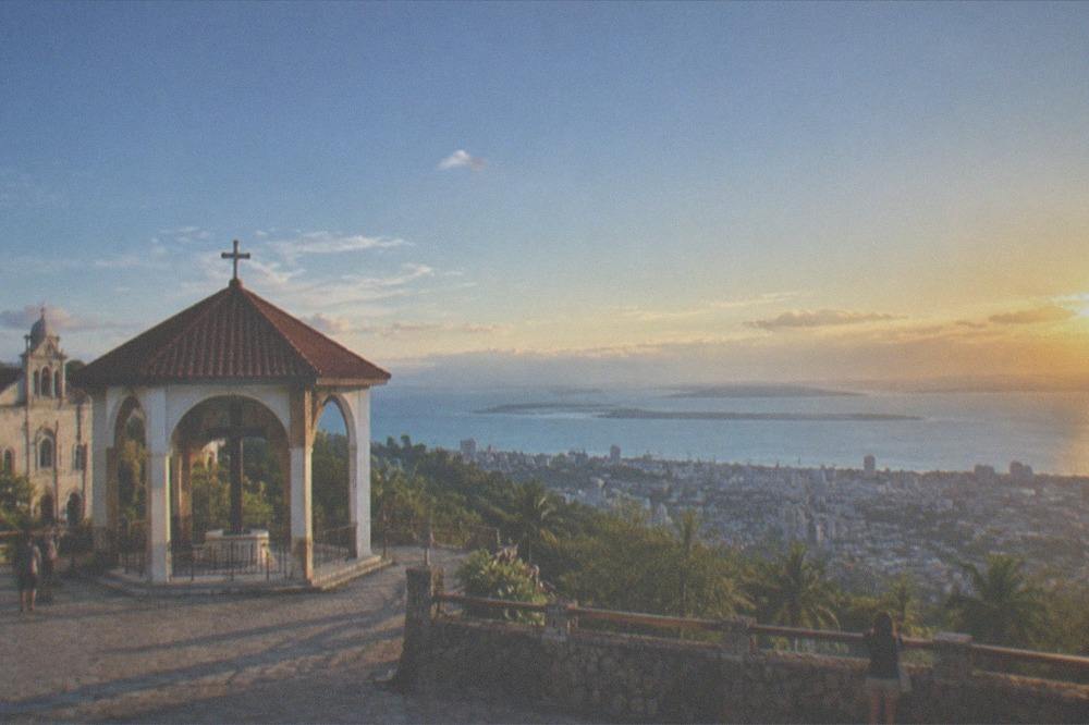 A mountain lookout platform above tropical hills at sunset with a city coastline below