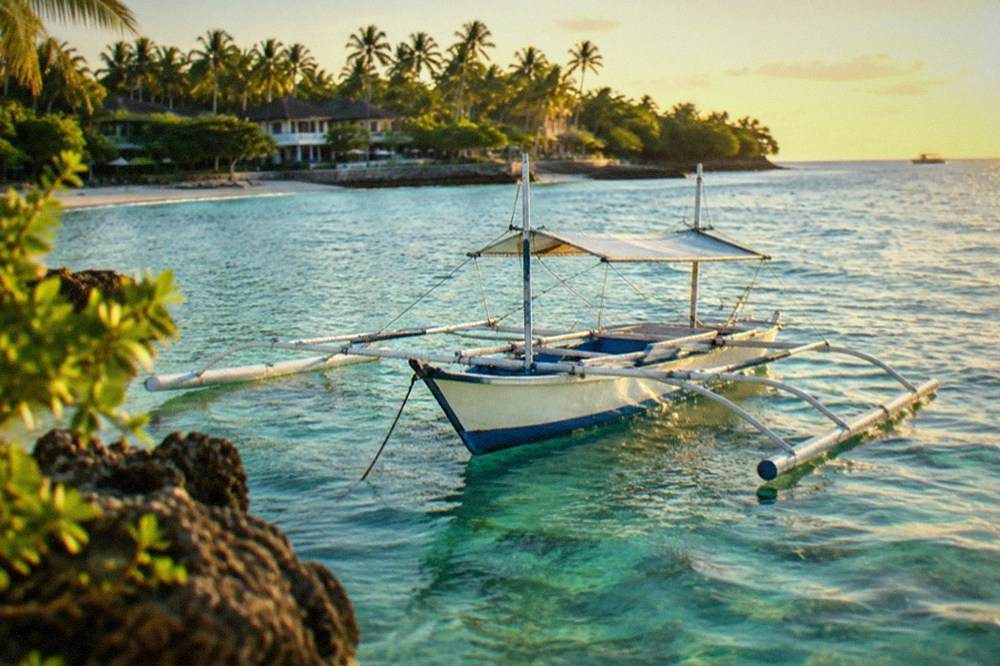 A small outrigger boat anchored over clear shallow water beside a tropical island near Mactan