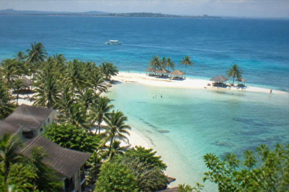 A white sandbar with a few palm-thatched shelters in shallow turquoise water in the Philippines