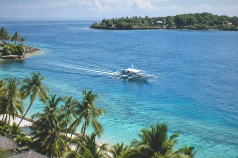 An outrigger banca boat crossing calm blue water toward a green tropical island near Mactan, Cebu
